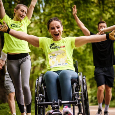 Bilde fra Wings for life world run. En kvinnelig utøver i rullestol har armene utover og smiler. Bak henne er det flere løpere med armene i været.
