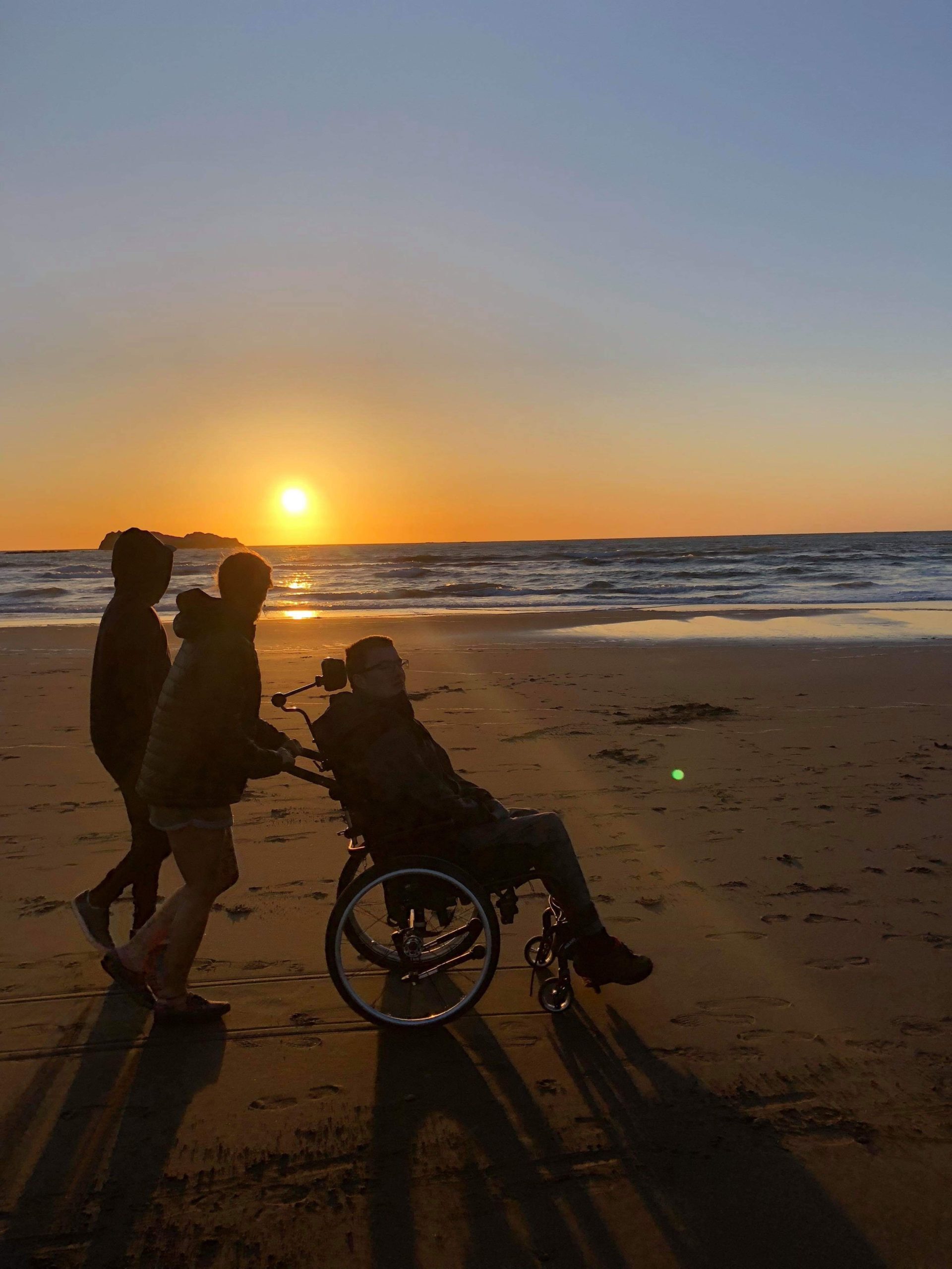 Fredrik trilles i rullestol på stranden av to assistenter i en nydelig solnedgang. Vi ser kun silhuettene av dem.