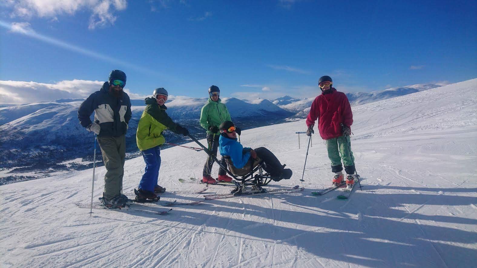 Fredrik på sitski med fire andre på fjellet. Flott landskap og blå himmel bak dem. Fargerike skiklær.