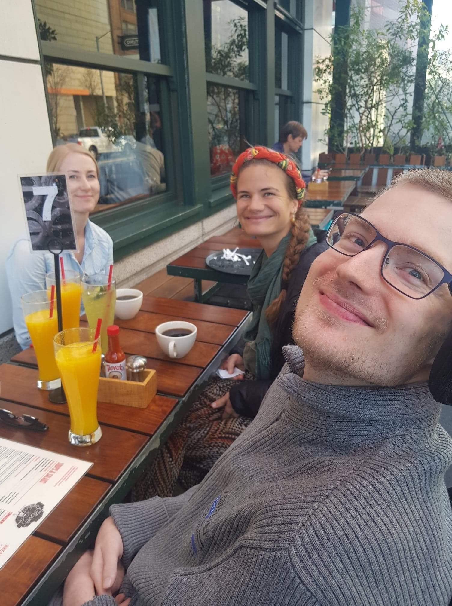 Fredrik sitter utendørs på restaurant med to kvinner. Alle ser inn i kamera og smiler. På bordet står det drinker.