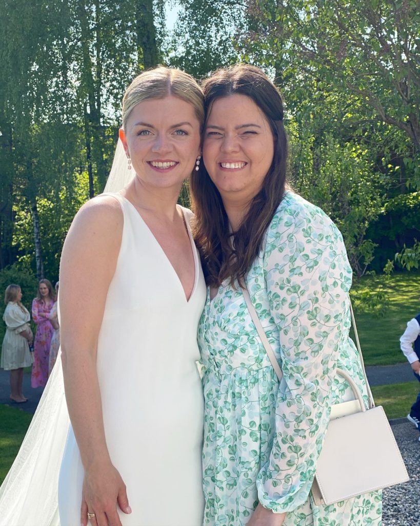 Tamarin sammen med venninnen som er brud. Bruden har på seg en hvit, klassisk ermeløs brudekjole og slør. Hun er blond. Tamarin har på lys og blå kjole med blå blomster. Hun har brunt hår. De står tett sammen og smiler til kamera.