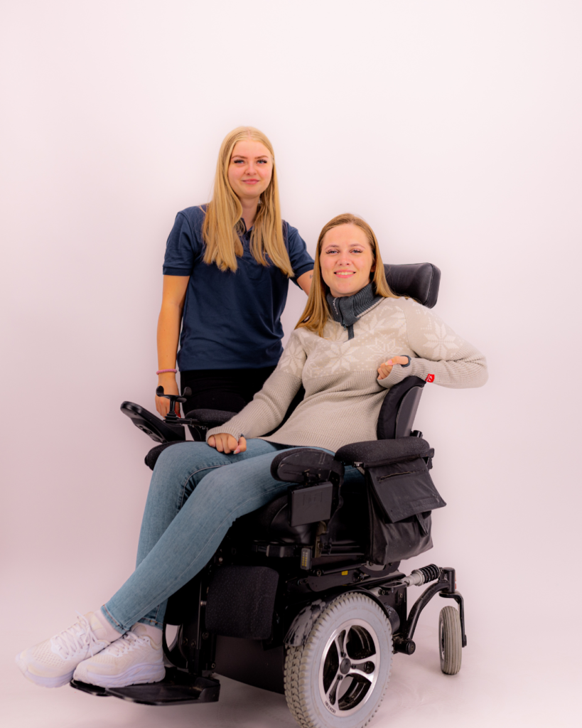 Hannah sammen med funksjonassistent Gina i fotostudio. Gina har blondt hår og har på seg en mørkeblå t-skjorte. Hannah har blondt hår og har på seg en lysegrå Ulvang-genser, lyseblå jeans og hvite sko. Hun sitter i en elektrisk rullestol. Begge smiler til kamera.