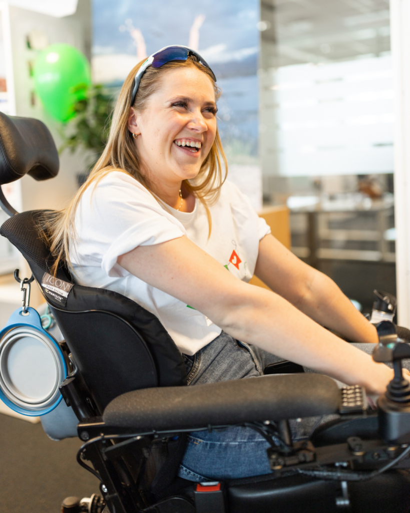 Hannah smiler stort. Hun har blondt hår, har på blå jeans, hvit t-skjorte og raske briller plassert på hodet. Hun sitter i elektrisk rullestol. Kontorlandskap i bakgrunnen.