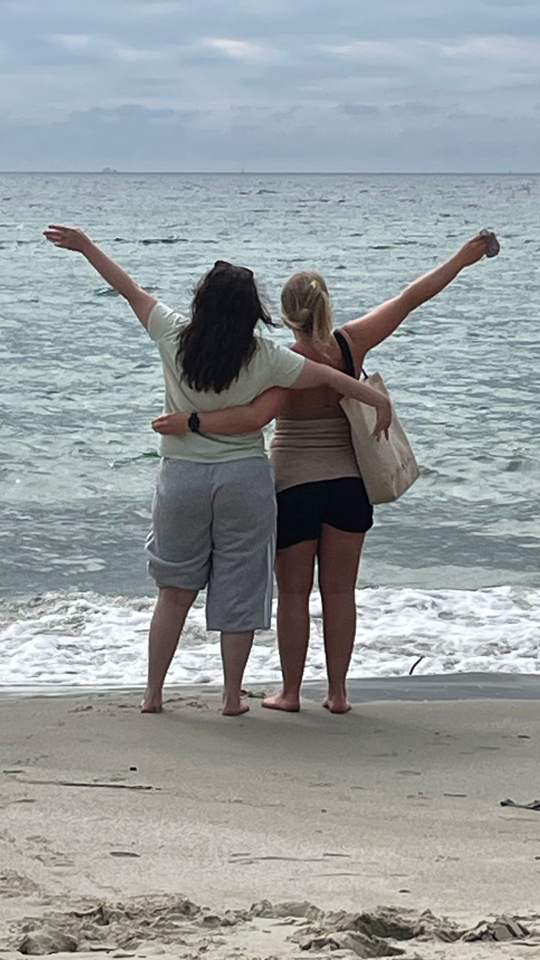 Tamarin med kvinnelig assistent på stranden. De holder rundt hverandre med den ene armen og har den andre i været. Vi ser dem bakfra. Bølger i havet.