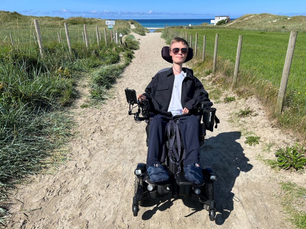 Fredrik foran sandveien ned til stranden og havet. Han sitter i elektrisk rullestol, har på seg svarte solbriller, svart jakke og mørkeblå bukser. Det er grønt gress ved siden av veien.