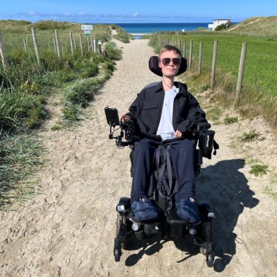 Fredrik foran sandveien ned til stranden og havet. Han sitter i elektrisk rullestol, har på seg svarte solbriller, svart jakke og mørkeblå bukser. Det er grønt gress ved siden av veien.