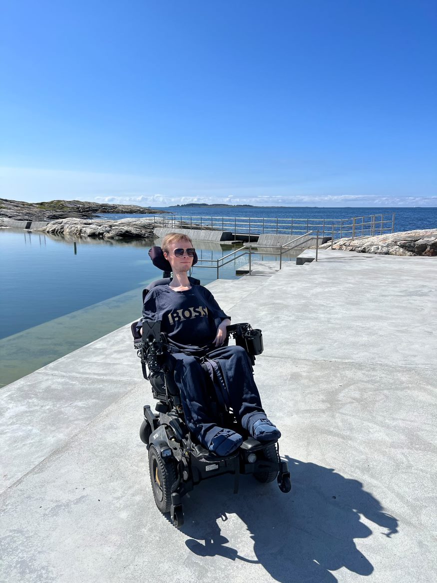 Fredrik i elektrisk rullestol foran Sjøbadet Myklebust. Svaberg i bakgrunnen, betongplatting og rullestolrampe.