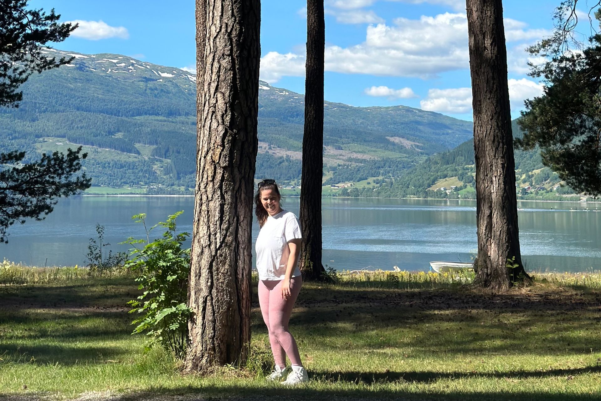 Tamarin, en ung dame, står ved siden av et høyt tre på grønt gress foran et vann og fjell i bakgrunnen. Har på rosa treningstights og hvit t-skjorte. Brunt, langt hår.
