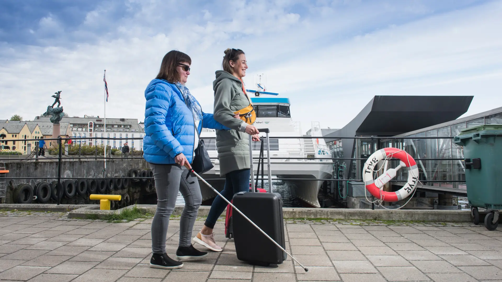 Kvinne med hvit stokk ledsages av kvinnelig assistent ved en fergehavn. Assistenten triller en koffert.