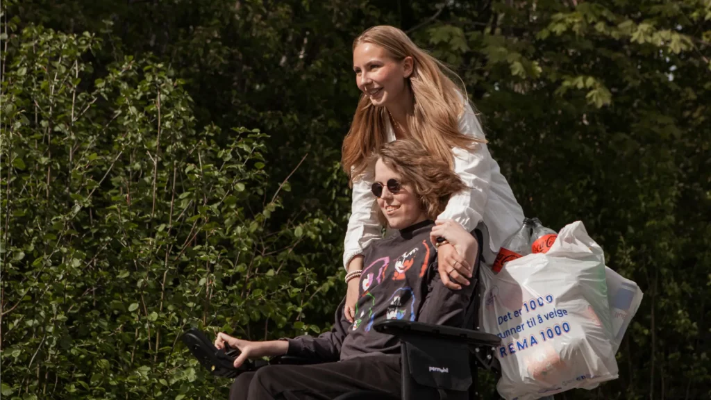 Jente i elektrisk rullestol med ung kvinnelig BPA-assistent stående bakpå. Begge smiler. Foran grønne busker.