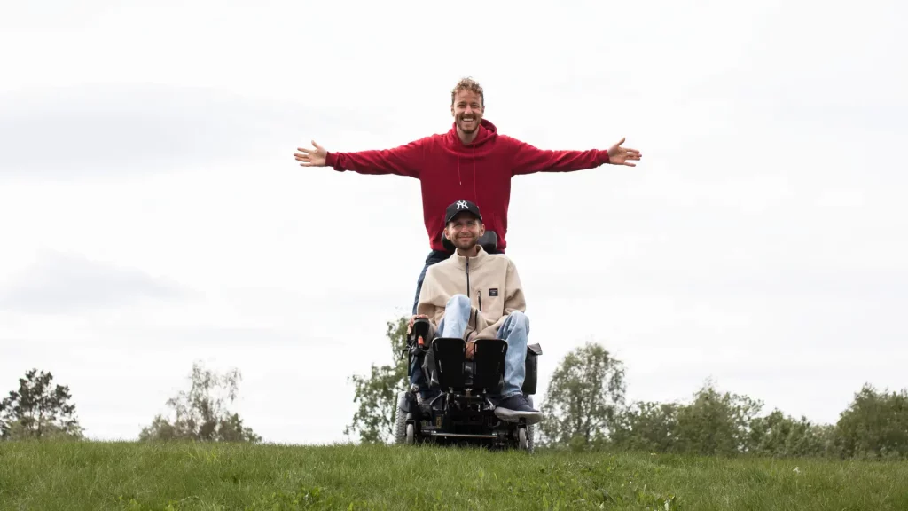 Mann i elektrisk rullestol på en gressplen med en mannlig BPA-assistent stående bak seg med utstrakte armer. Begge smiler.