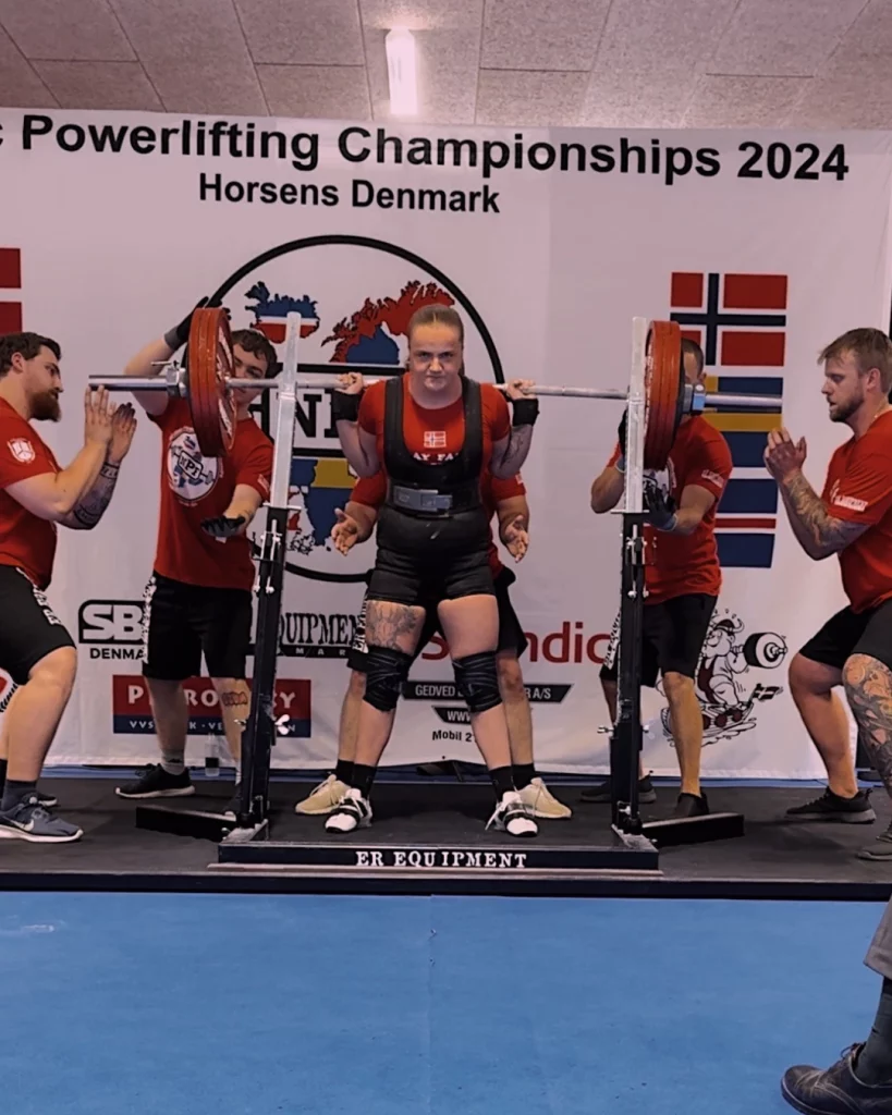 Ung kvinne løfter stang med vekter over skuldrene. Flere personer står rundt henne for å assistere. På veggen bak står det "Powerlifting championships 2024".