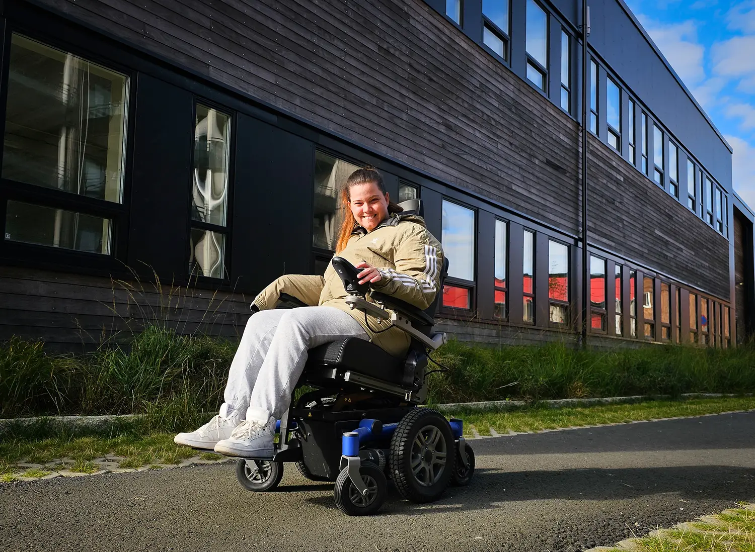 Ung kvinne i elektrisk rullestol utenfor en boligblokk. Grønn jakke, grå bukser. Hun smiler.