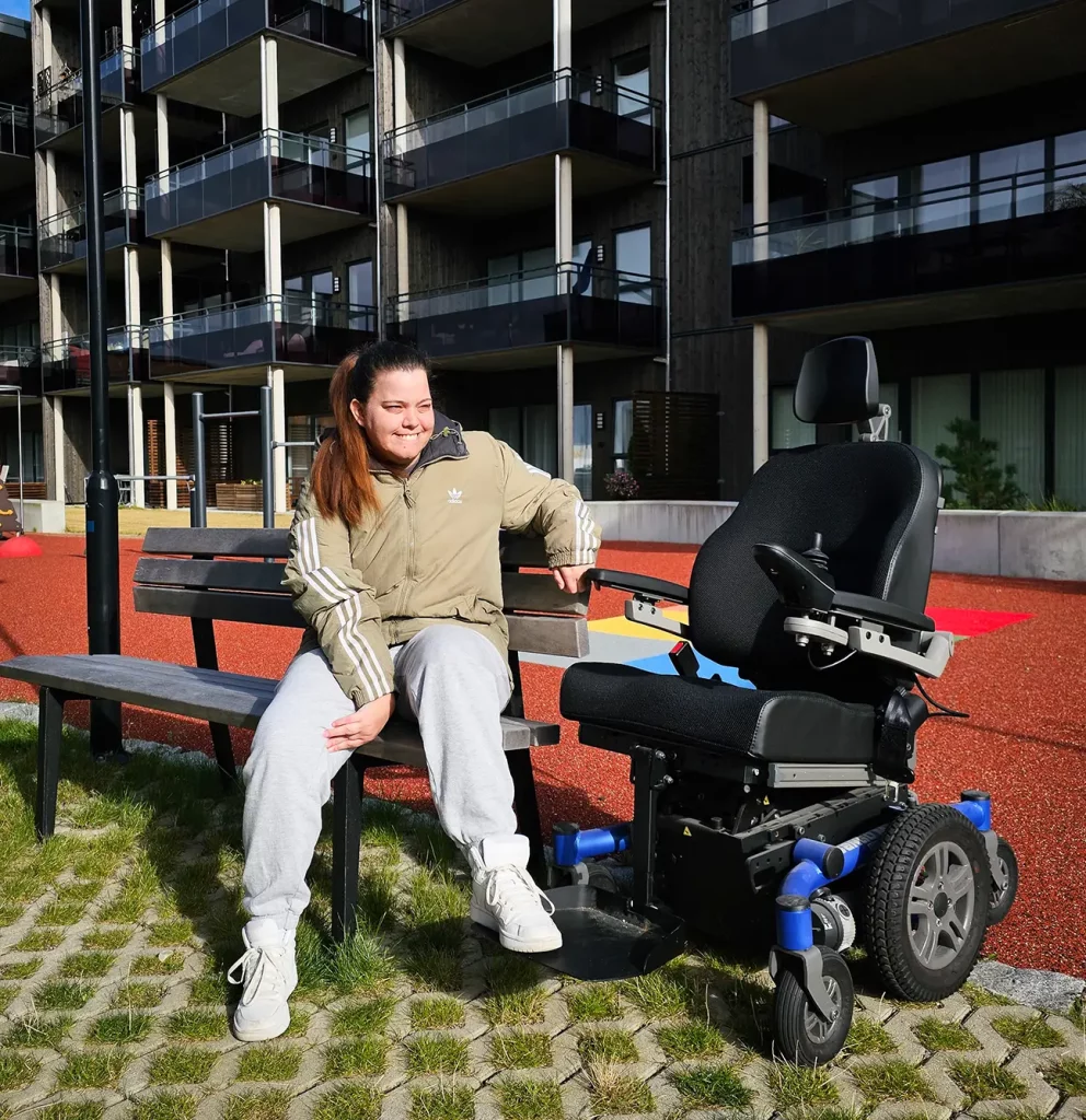 Kvinne med brunt hår i hestehale, grønn jakke og grå bukser sitter på en benk. Ved siden av står en elektrisk rullestol. 