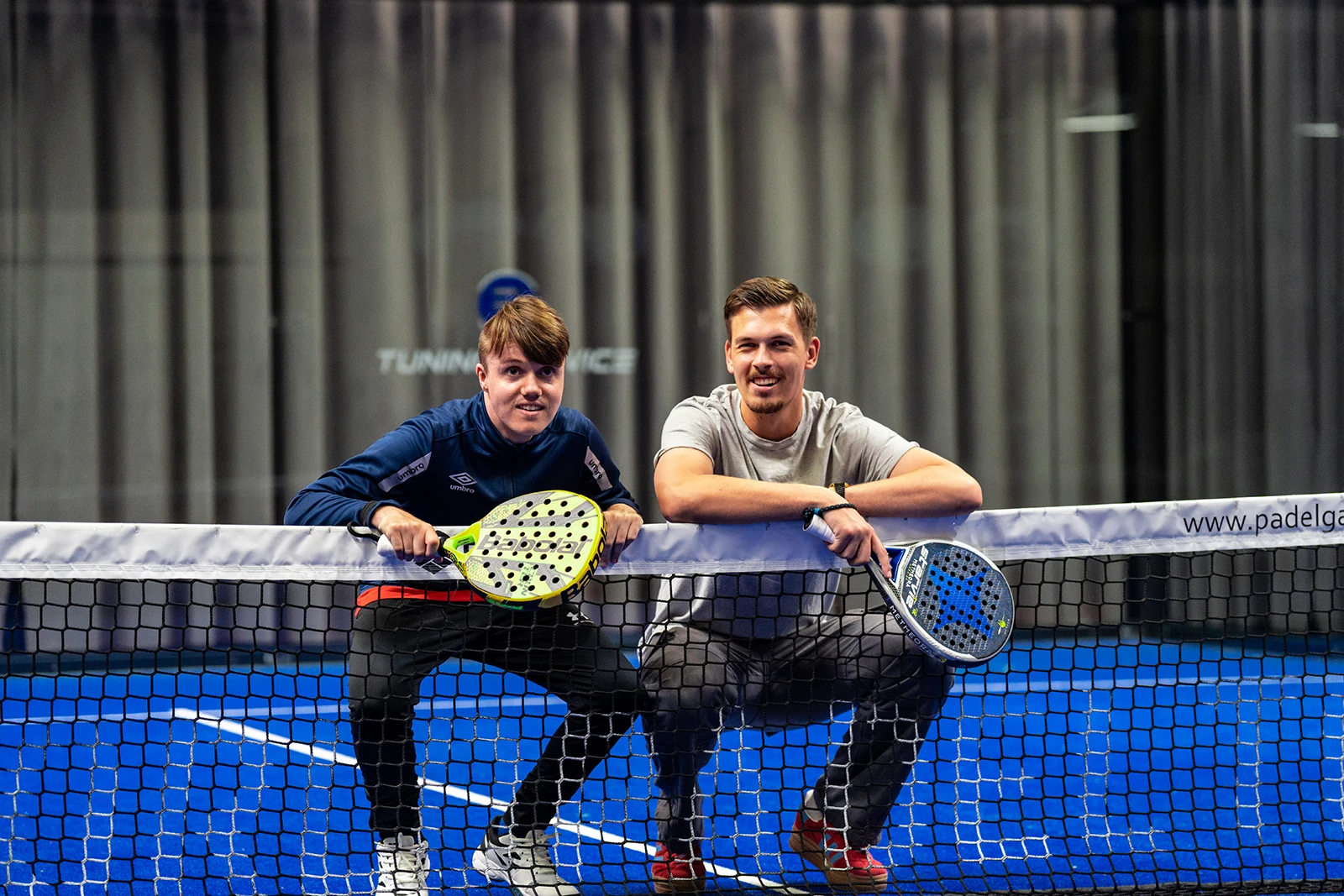 Mannlig assistent sammen med ung mann. begge smiler og ser inn i kamera. De befinner seg på en Padel bane.