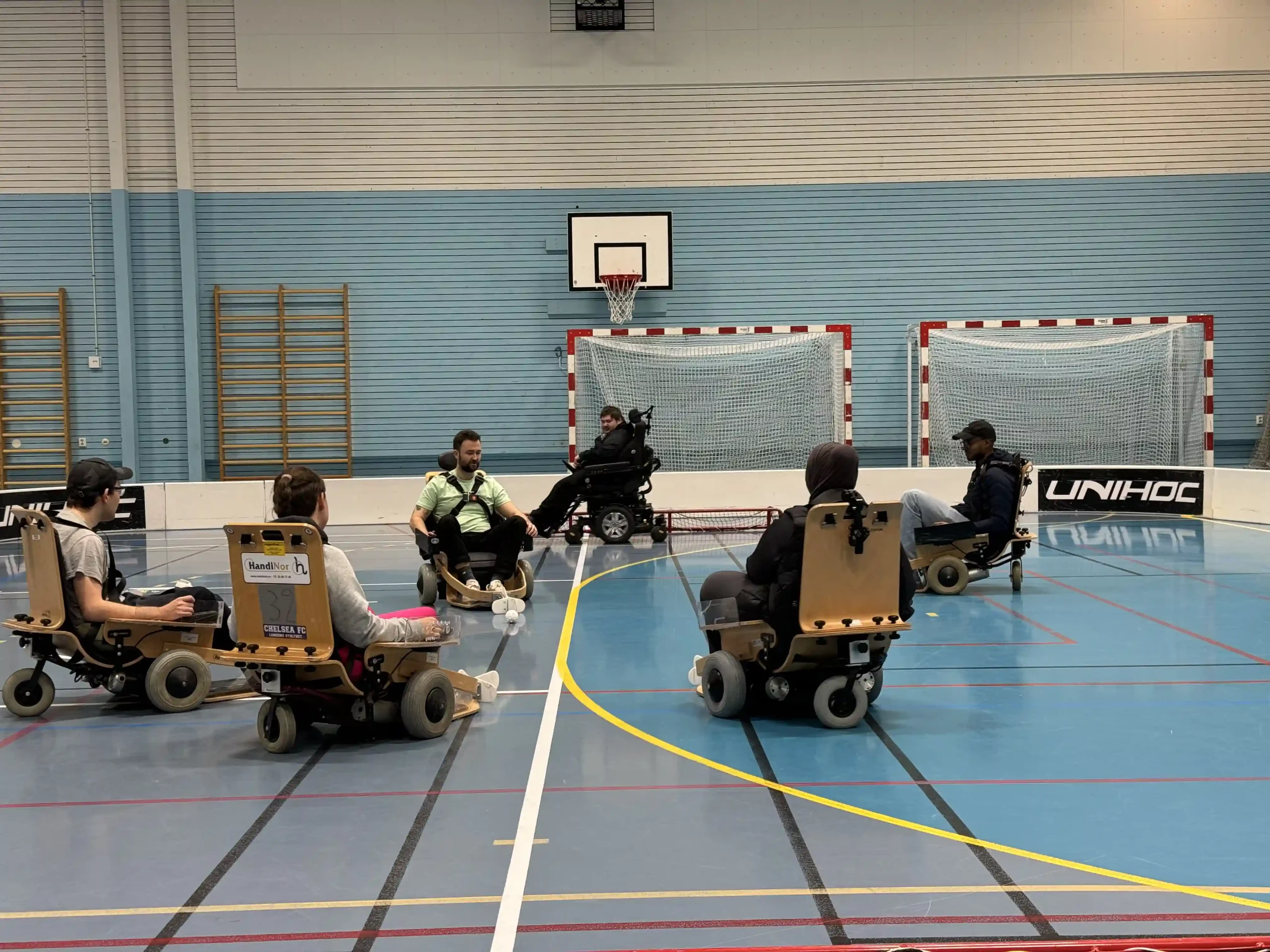 6 personer som sitter i El-innebandy stoler inne i idrettshall