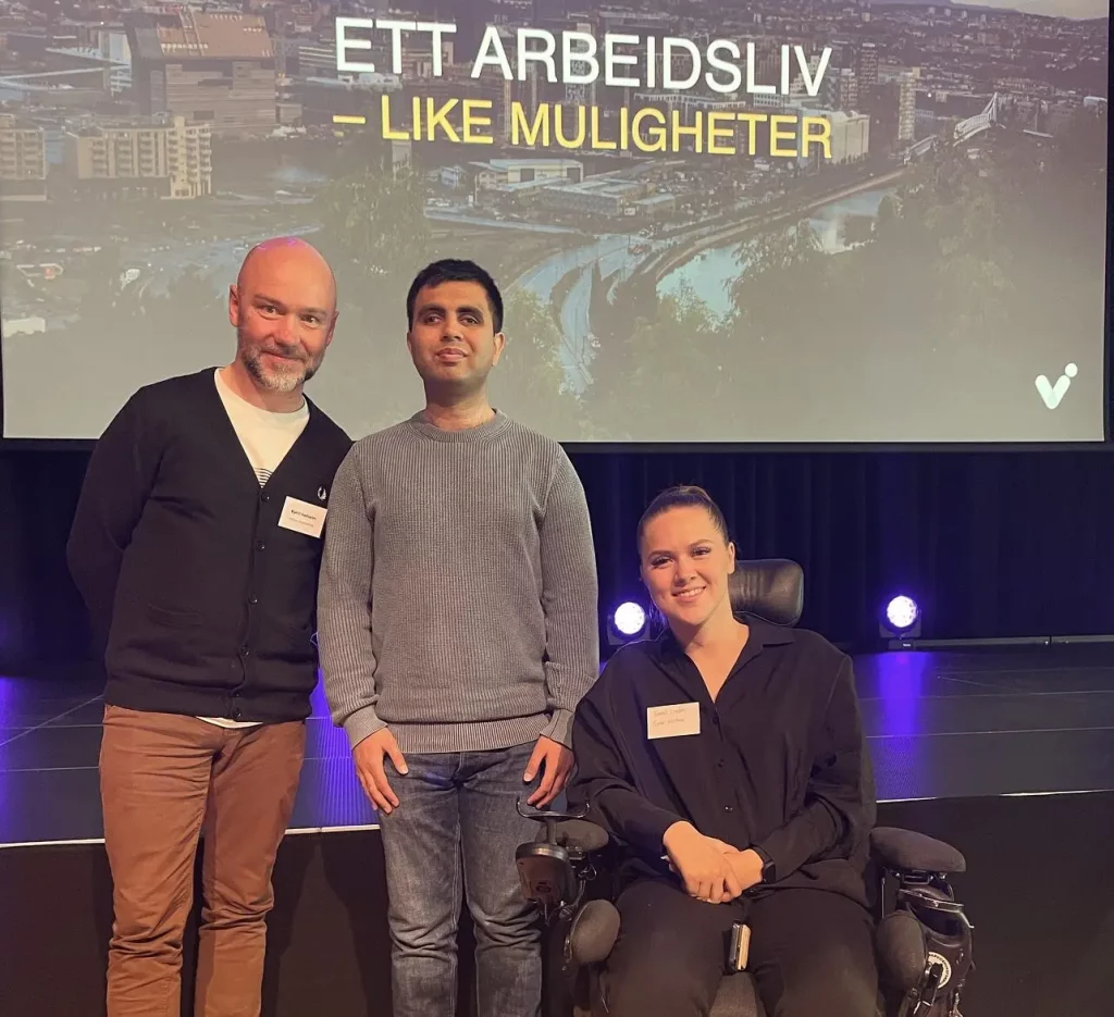 Atif, sjef Kjetil og kollega Hannah smiler til kamera og står foran en stor skjerm hvor det står "Ett arbeidsliv - like muligheter". Hannah sitter i rullestol. 