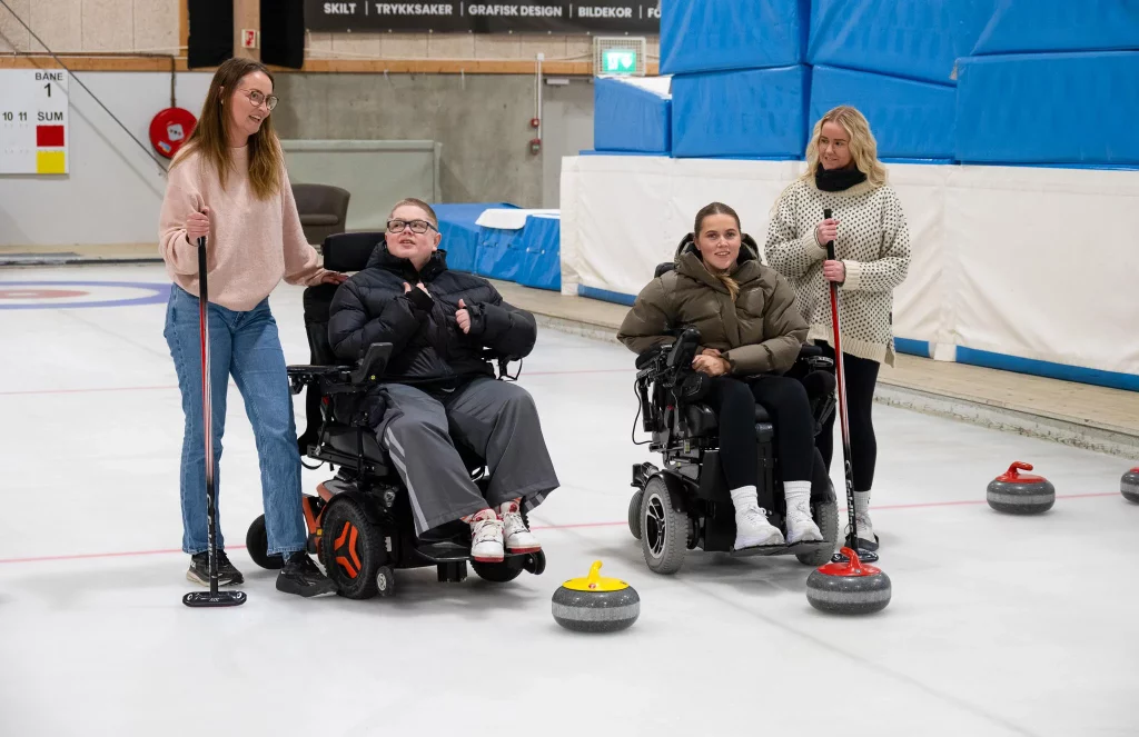 ei jente og en gutt i rullestol sammen med to kvinnelige BPA assistenter på curlingbane