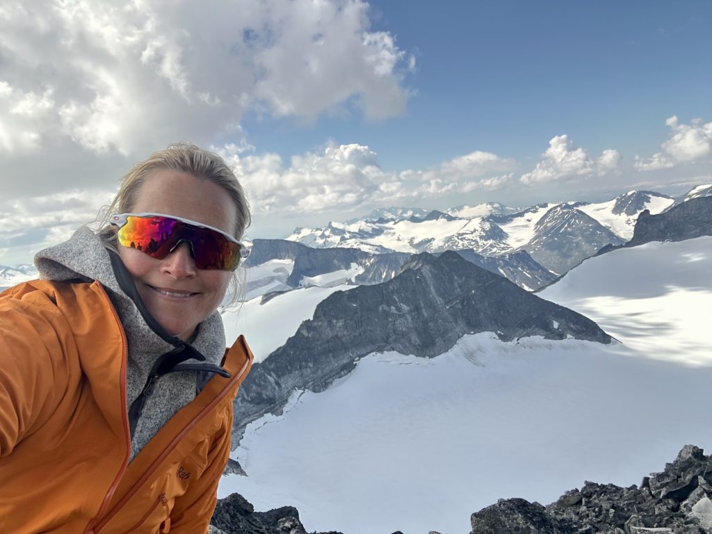 dame som smiler mot kamera med utsikt over snøkledde fjell i bakgrunnen
