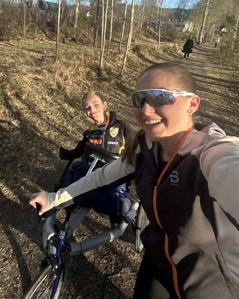 Ung kvinne tar en selfie på en gangsti mellom trær. Hun har på raske briller og holder i den tilpassede sykkelen til en ung gutt som smiler inn i kamera.