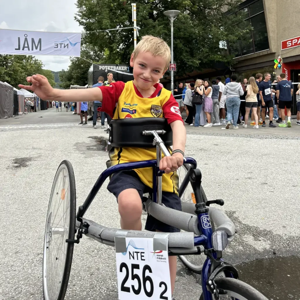 gutt som smiler mot kamera ved målstrek. gutten sitter i en frame running stol