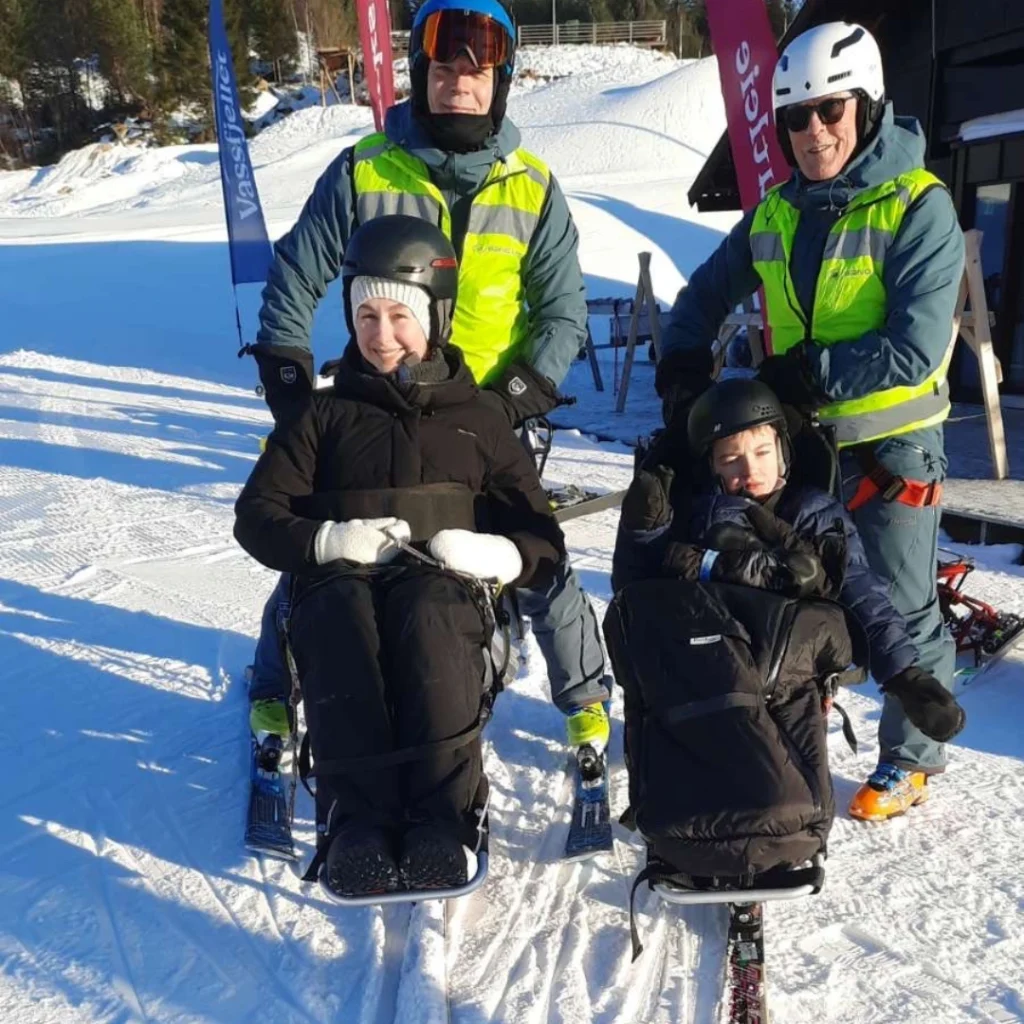 Ung gutt og ung jente i slalombakke sittende i sitski. Bak dem står to menn med refleksvester.